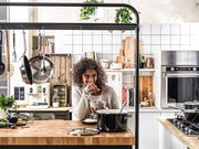 Frau in moderner Küche mit einem schwarzen Topfset auf dem Herd, fröhlich kochend. Perspektive: Frontalansicht.