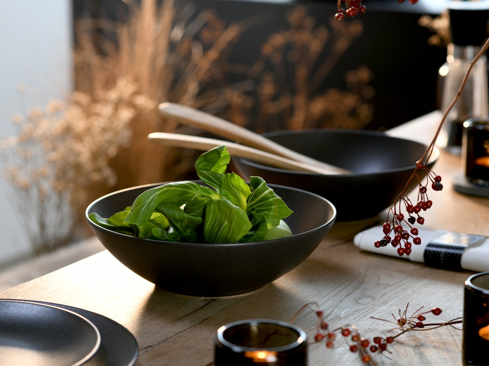 Creatable Salatschüssel 2-tlg SOFT TOUCH Mattschwarze Keramikschale mit frischen grünen Blättern auf einem Holztisch, aufgenommen in schräger Seitenansicht mit leicht erhöhter Perspektive; im Hintergrund weitere Schalen und Holzlöffel.