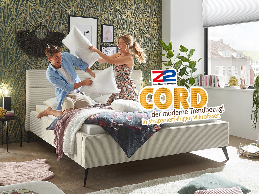 Junges Paar bei einer fröhlichen Kissenschlacht auf einem modernen Polsterbett mit hellem Cordbezug in einem stilvoll eingerichteten Schlafzimmer mit trendiger Tapete und Pflanzen.