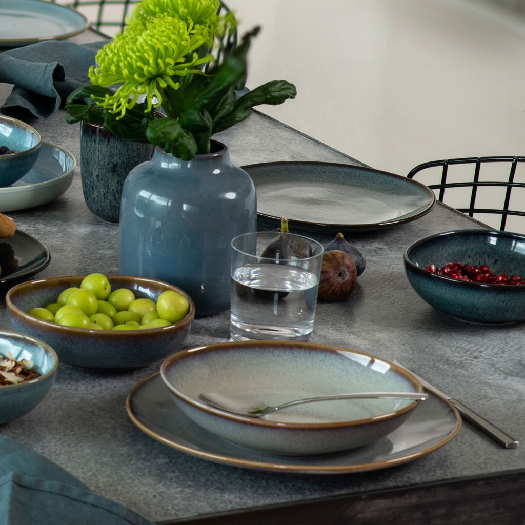 Speiseteller Lave 28cm türkis auf einem gedeckten Tisch, von oben betrachtet, umgeben von Schalen mit Obst und einer Vase mit Blumen.