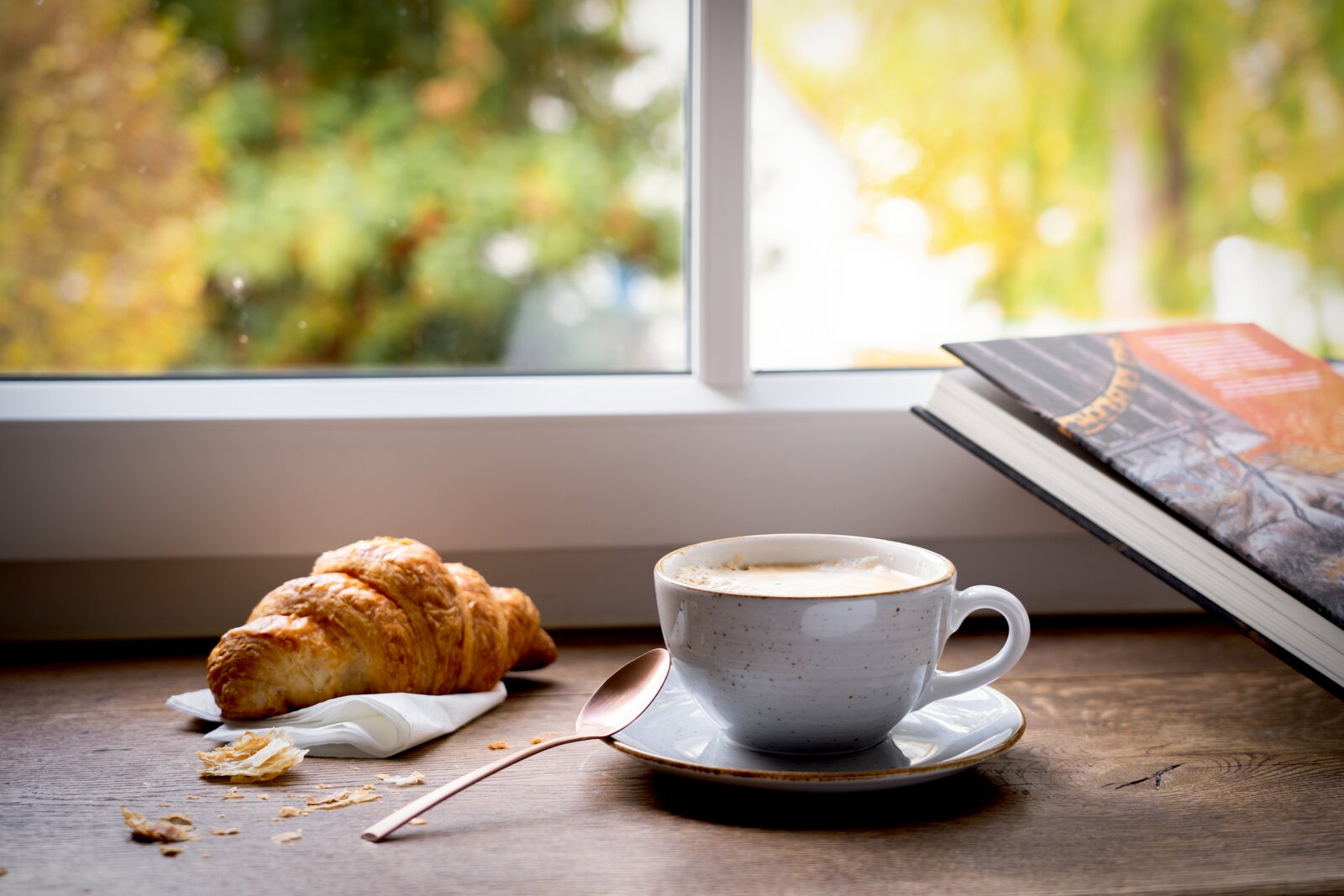 Weiße Keramik-Kaffeetasse mit Milchkaffee auf Untertasse neben Croissant und Löffel auf Holztisch am Fenster, perspektivisch aus Augenhöhe in leichter Seitenansicht aufgenommen