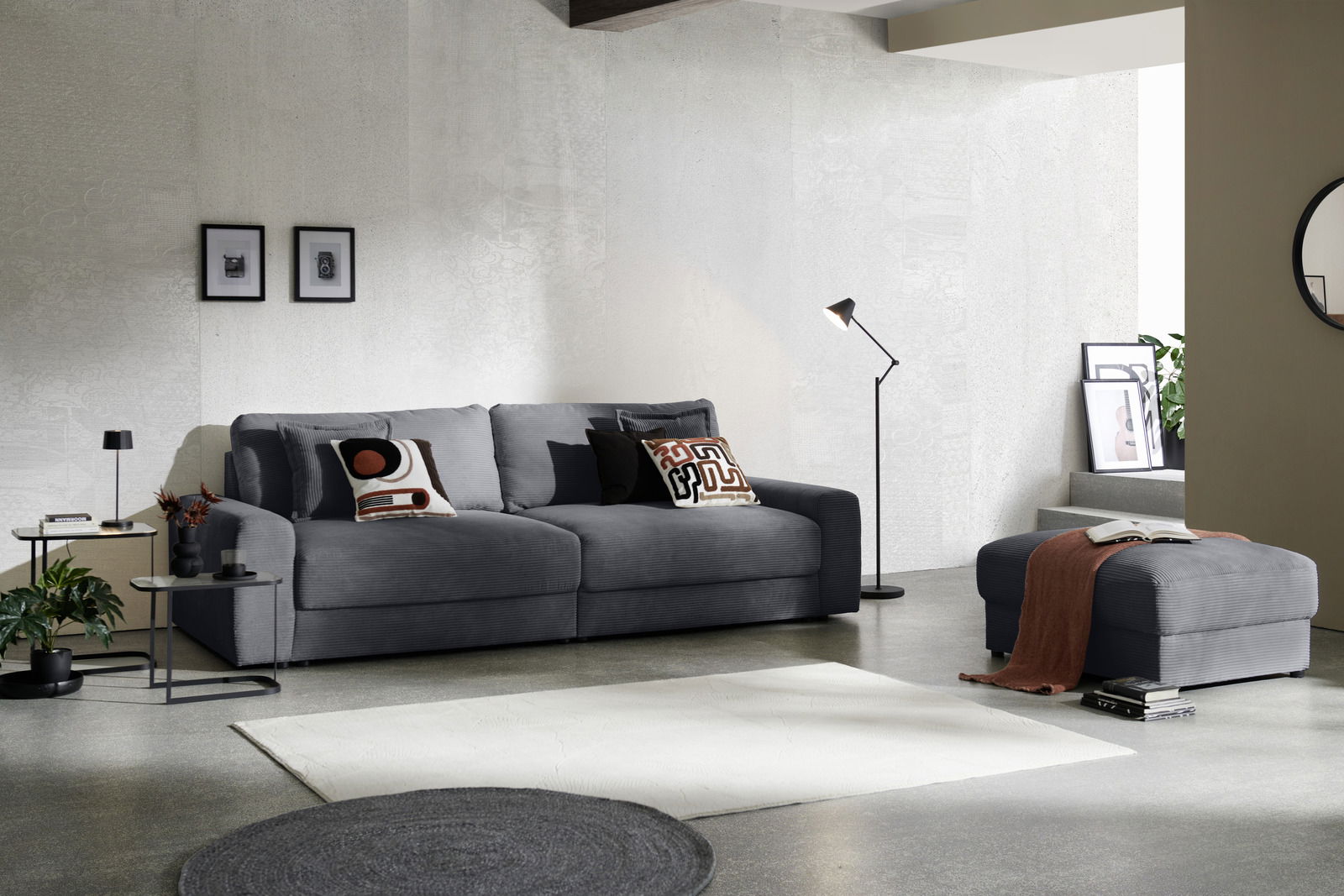 Großes, graues Big-Sofa in modernem Wohnzimmer, seitliche Perspektive, mit dekorativen Kissen und passendem Hocker.