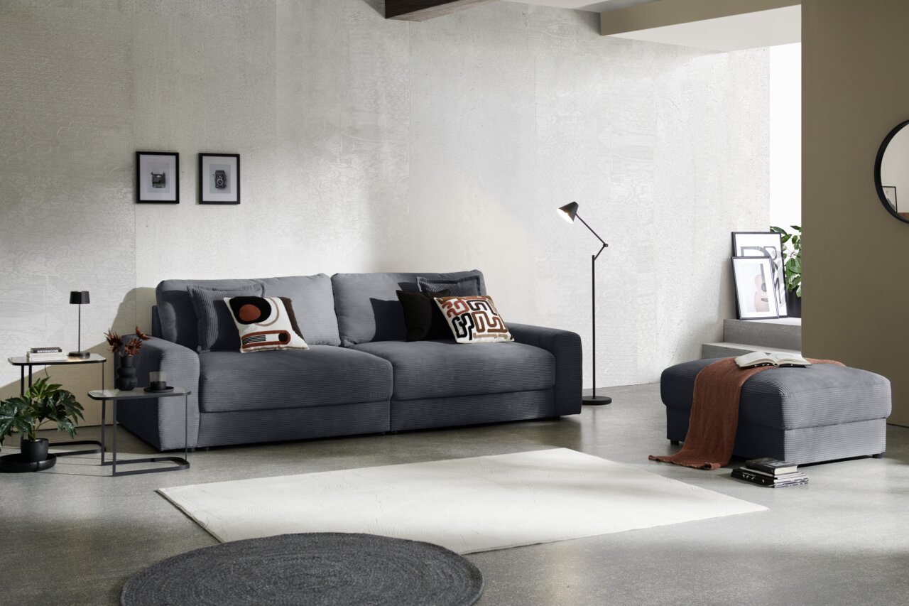 Z2 Big Sofa NAPOLI Großes, graues Big-Sofa in modernem Wohnzimmer, seitliche Perspektive, mit dekorativen Kissen und passendem Hocker.