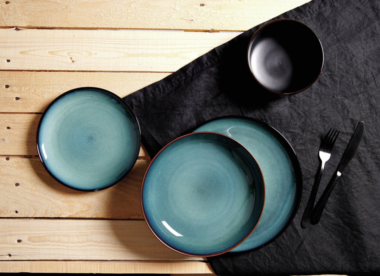 Vogelperspektive auf ein 8-teiliges Tafelservice mit blauen Tellern und einer schwarzen Schüssel auf einem Holztisch mit schwarzer Tischdecke.