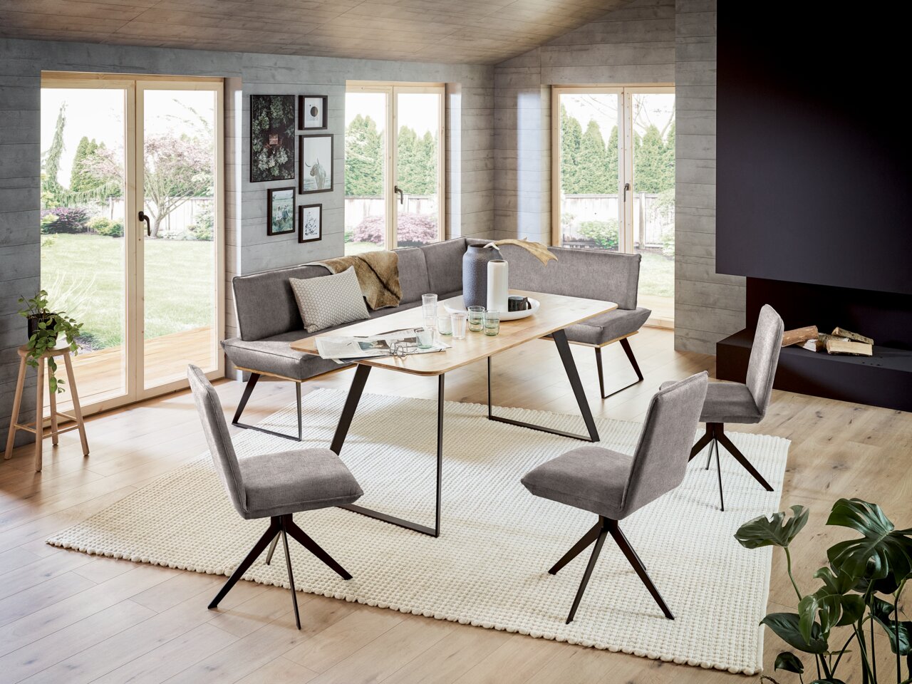 Voleo Eckbank MALIBU Moderne Esszimmer-Eckbank mit grauen Polstern und schwarzem Metallgestell, umgeben von passenden Stühlen und einem Holztisch, in einem hellen Raum mit großen Fenstern und Blick auf den Garten. Perspektive: seitlich von vorne.