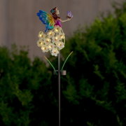 Verlichte tuindecoratie in de vorm van een spies met een elfje, kleurrijke vlindervleugels en lichtgevende bloemen-leds op een metalen staaf, van voren gefotografeerd vanuit een licht zijdelings perspectief