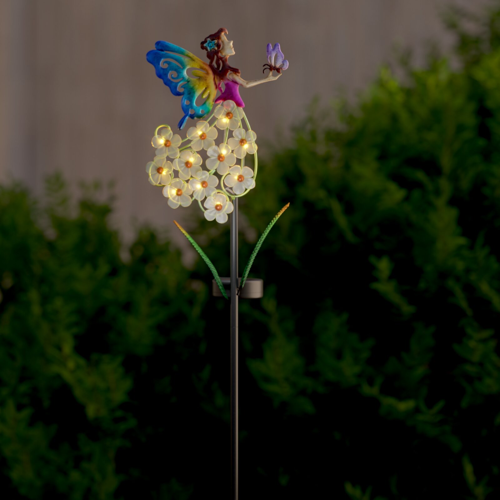 Verlichte tuindecoratie in de vorm van een spies met een elfje, kleurrijke vlindervleugels en lichtgevende bloemen-leds op een metalen staaf, van voren gefotografeerd vanuit een licht zijdelings perspectief