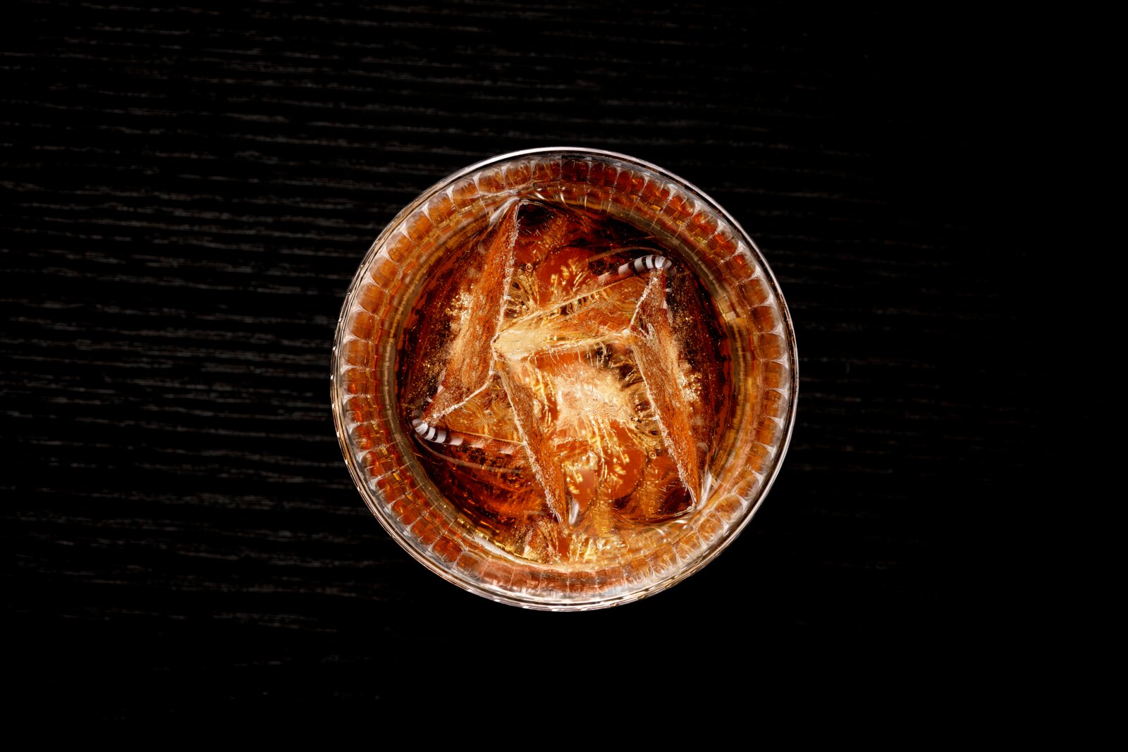 Bovenaanzicht van een glas met een drankje en ijsblokjes op een donkere tafel.