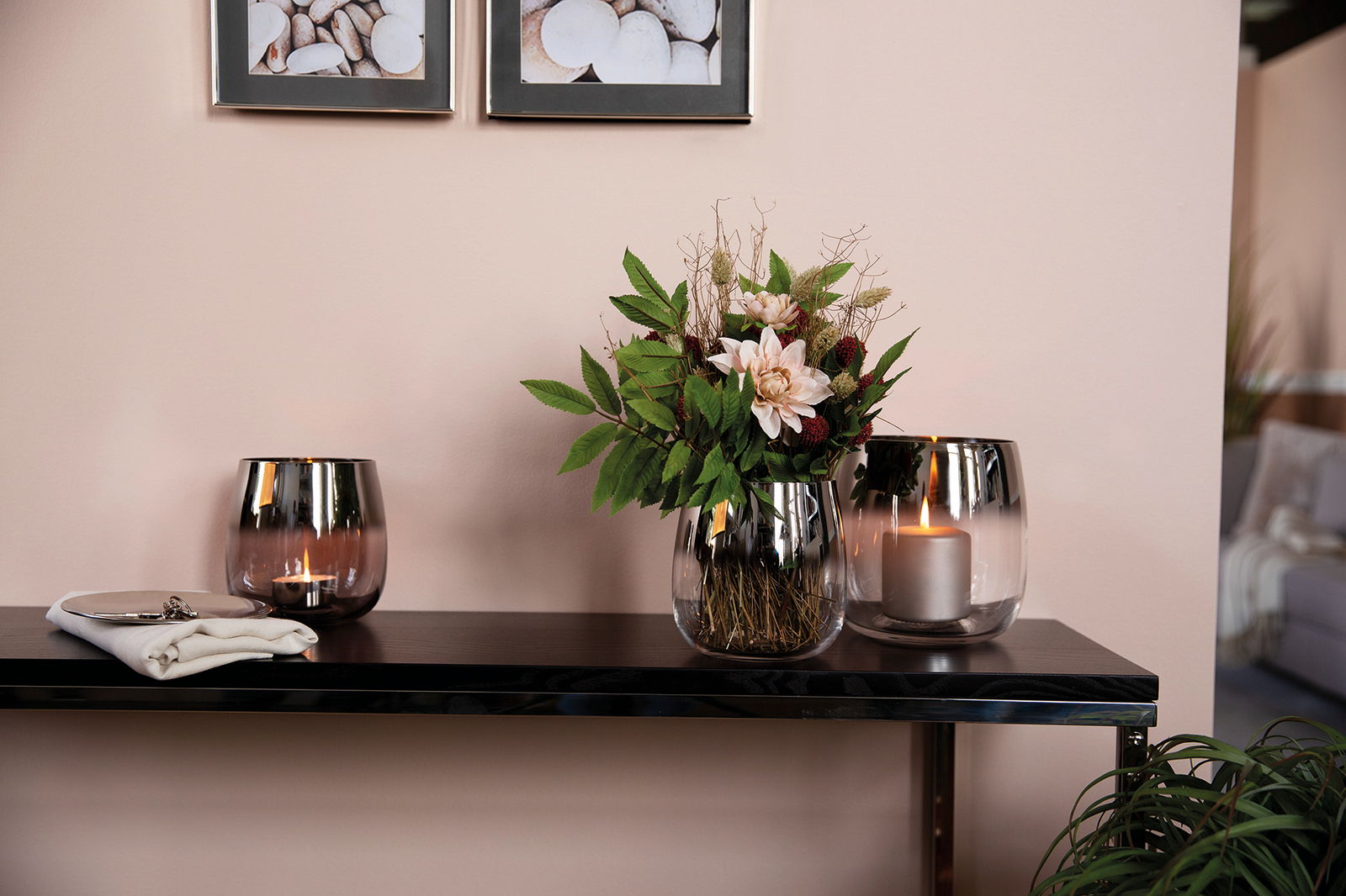 Drei klare Glasvasen auf einem Regal, eine mit Blumenarrangement, die anderen mit Kerzen, aus seitlicher Perspektive.