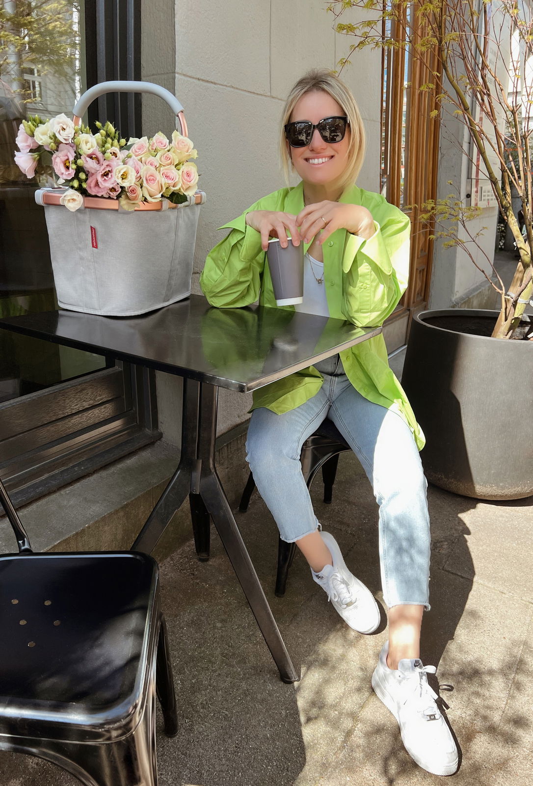 Eine Frau sitzt an einem Tisch im Freien und genießt ein Getränk. Neben ihr steht ein grauer Einkaufskorb mit Blumen, der von der Seite zu sehen ist.