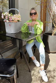 Eine Frau sitzt an einem Tisch im Freien und genießt ein Getränk. Neben ihr steht ein grauer Einkaufskorb mit Blumen, der von der Seite zu sehen ist.