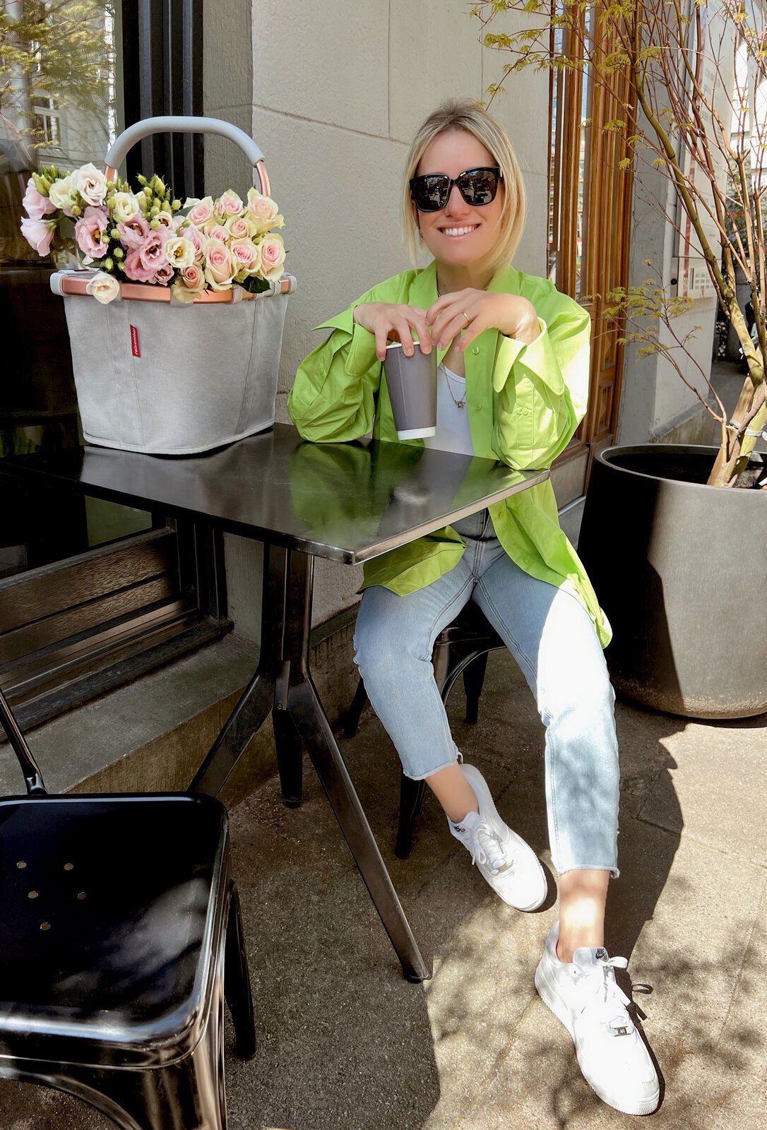 Reisenthel Carrybag Twist Sky Rose Eine Frau sitzt an einem Tisch im Freien und genießt ein Getränk. Neben ihr steht ein grauer Einkaufskorb mit Blumen, der von der Seite zu sehen ist.