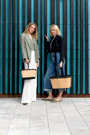 Zwei Frauen stehen vor einer modernen Wand und halten jeweils eine beige carrybag XS mit schwarzem Griff. Die Perspektive ist frontal.