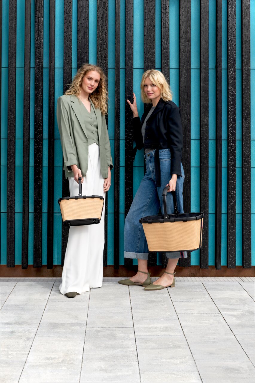 Zwei Frauen stehen vor einer modernen Wand und halten jeweils eine beige carrybag XS mit schwarzem Griff. Die Perspektive ist frontal.