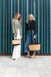 Zwei Frauen stehen vor einer modernen Wand und halten jeweils eine beige carrybag XS mit schwarzem Griff. Die Perspektive ist frontal.