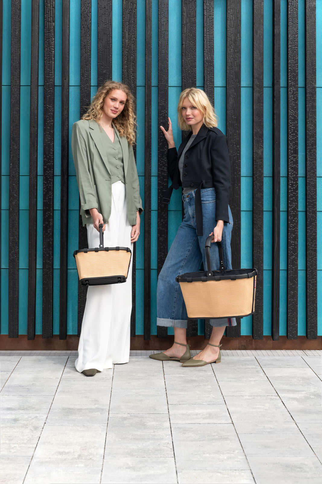 Zwei Frauen stehen vor einer modernen Wand und halten jeweils eine beige carrybag XS mit schwarzem Griff. Die Perspektive ist frontal.