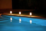 Drei leuchtende Akku-Tischleuchten MAIA am Rand eines Pools bei Nacht, mit Spiegelung im Wasser, seitliche Perspektive.