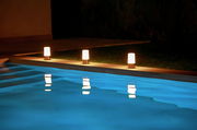 Drei leuchtende Akku-Tischleuchten MAIA am Rand eines Pools bei Nacht, mit Spiegelung im Wasser, seitliche Perspektive.