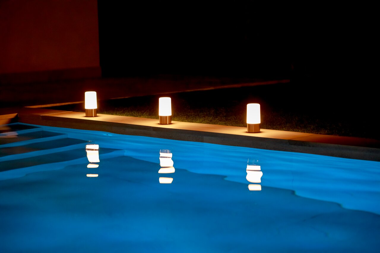 Drei leuchtende Akku-Tischleuchten MAIA am Rand eines Pools bei Nacht, mit Spiegelung im Wasser, seitliche Perspektive.