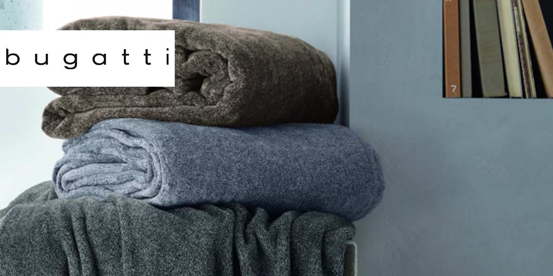 Drei zusammengerollte Kuscheldecken in Grau, Braun und Dunkelgrau, gestapelt vor einer hellblauen Wand, mit 