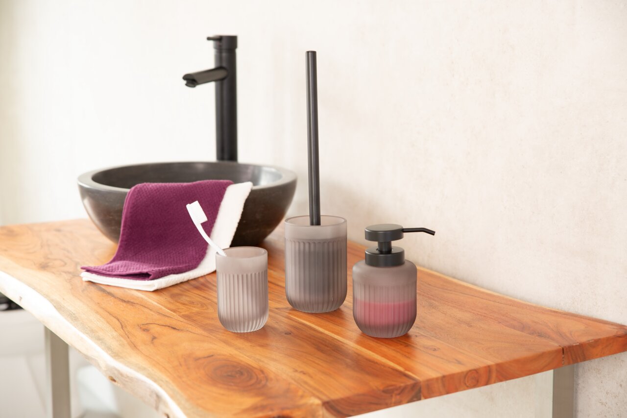 Sadena Zahnputzbecher VERA Drei graue Aufbewahrungsbecher Vera aus Glas mit geriffelter Oberfläche auf einem Holzwaschtisch, fotografiert aus seitlicher Perspektive. Im Hintergrund ein schwarzes Waschbecken und ein lila Handtuch.