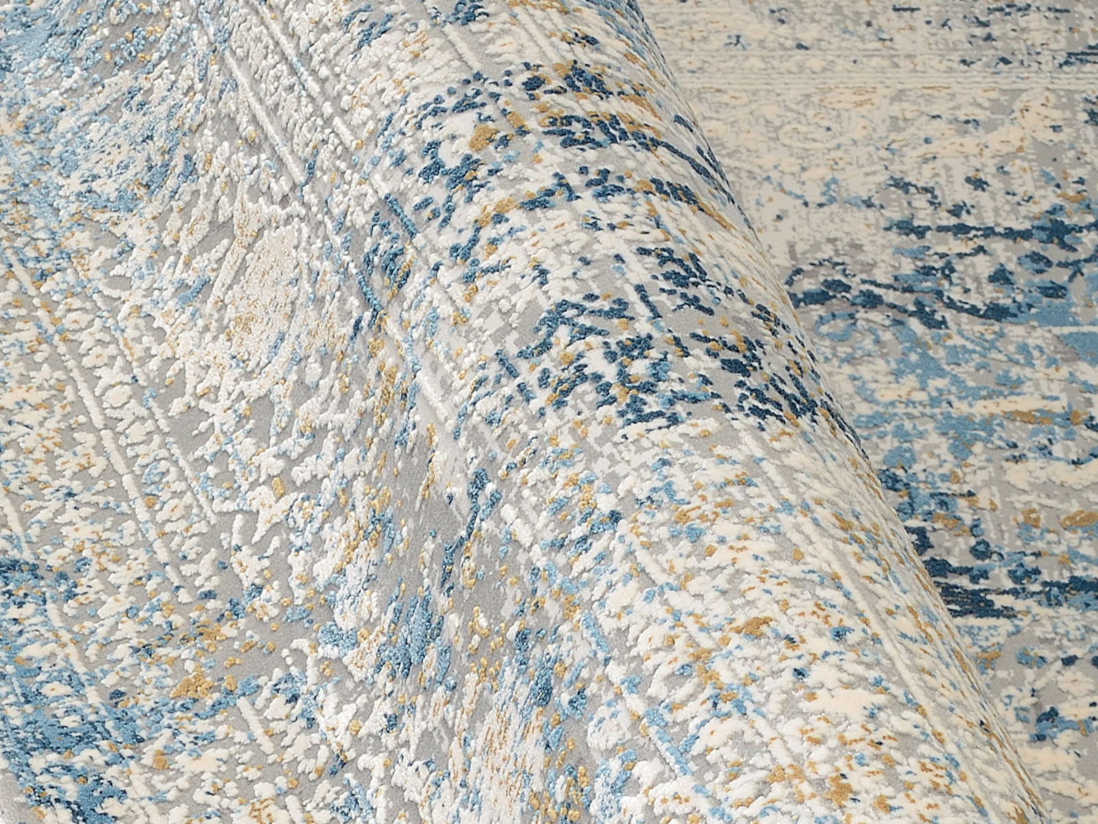 Nahaufnahme eines Designer-Teppichs mit abstraktem Muster in Blau-, Grau- und Beigetönen, leicht aufgerollt.