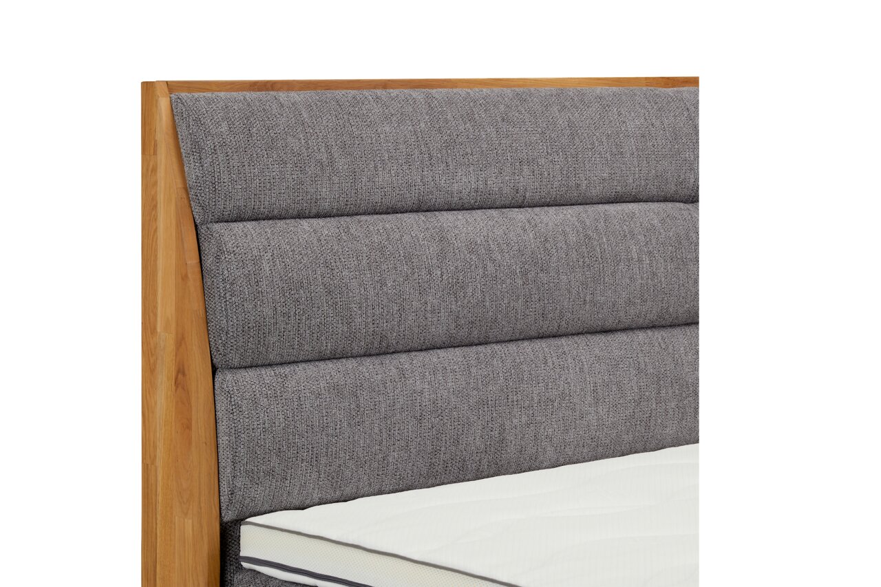 Close-up van een boxspringbed met een grijs, gestoffeerd hoofdeinde en een houten frame, gezien vanaf de zijkant