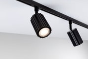 Schwarzer LED-Schienenstrahler mit geriffeltem zylindrischem Gehäuse an Deckenstromschiene, eingeschaltet, aus schräger Untersicht aufgenommen