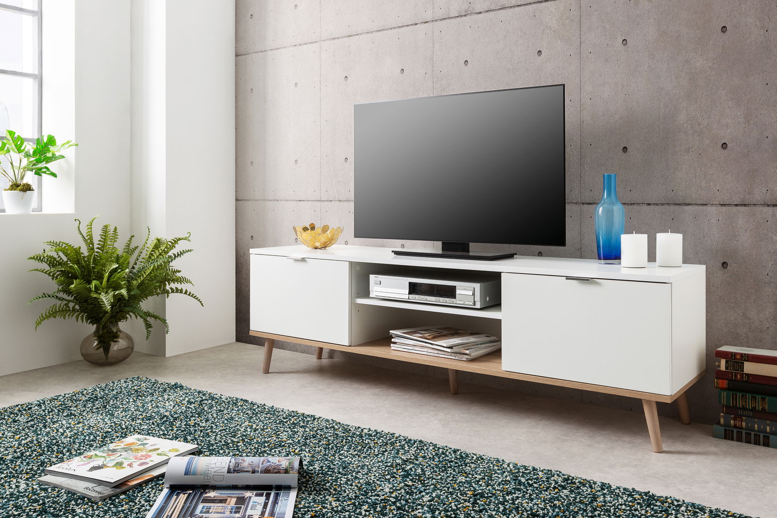 Modernes weißes TV-Lowboard mit Holzbeinen, dekoriert mit blauen und weißen Accessoires, in einem stilvollen Wohnzimmer mit Betonwand. Perspektive von vorne.