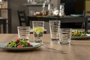 Vier Gläser mit strukturiertem Design auf einem Holztisch, gefüllt mit Wasser und Limettenscheiben, im Hintergrund ein gedeckter Tisch.