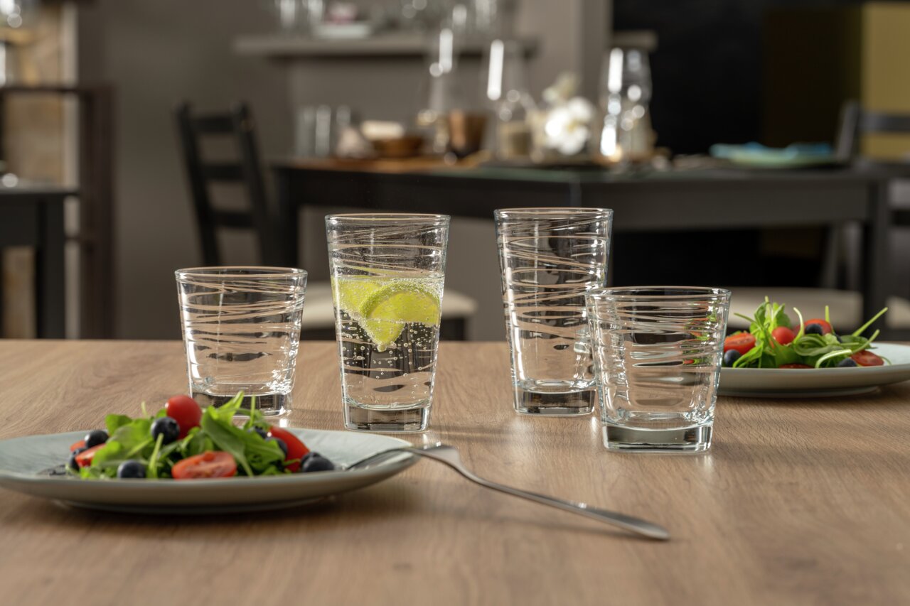 Vier Gläser mit strukturiertem Design auf einem Holztisch, gefüllt mit Wasser und Limettenscheiben, im Hintergrund ein gedeckter Tisch.