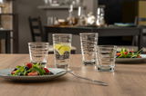 Vier Gläser mit strukturiertem Design auf einem Holztisch, gefüllt mit Wasser und Limettenscheiben, im Hintergrund ein gedeckter Tisch.