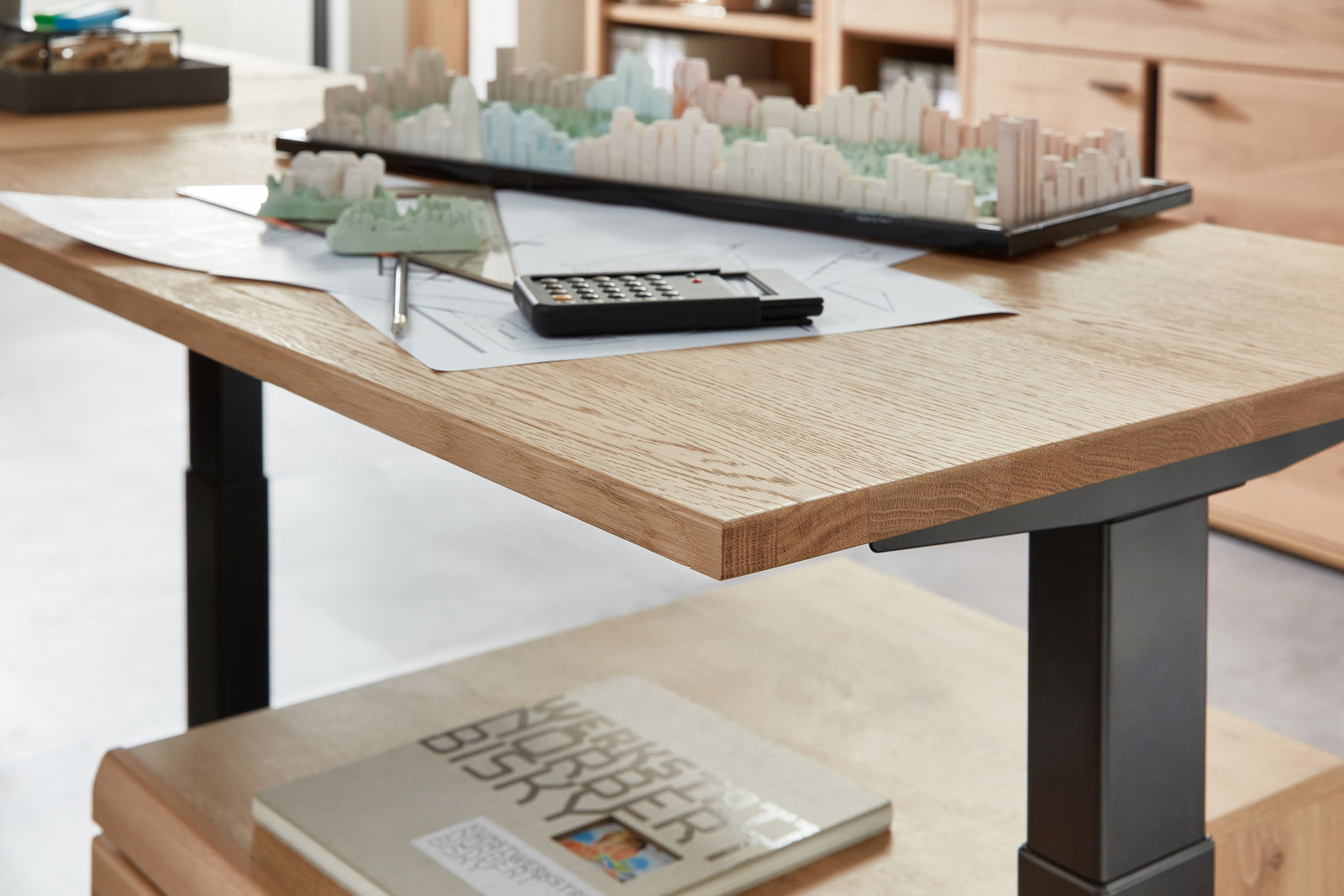 Nahaufnahme eines E-Schreibtischs mit Furnieroberfläche, auf dem ein Architekturmodell, ein Taschenrechner und Zeichnungen liegen. Perspektive von der Seite.