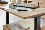 Nahaufnahme eines E-Schreibtischs mit Furnieroberfläche, auf dem ein Architekturmodell, ein Taschenrechner und Zeichnungen liegen. Perspektive von der Seite.
