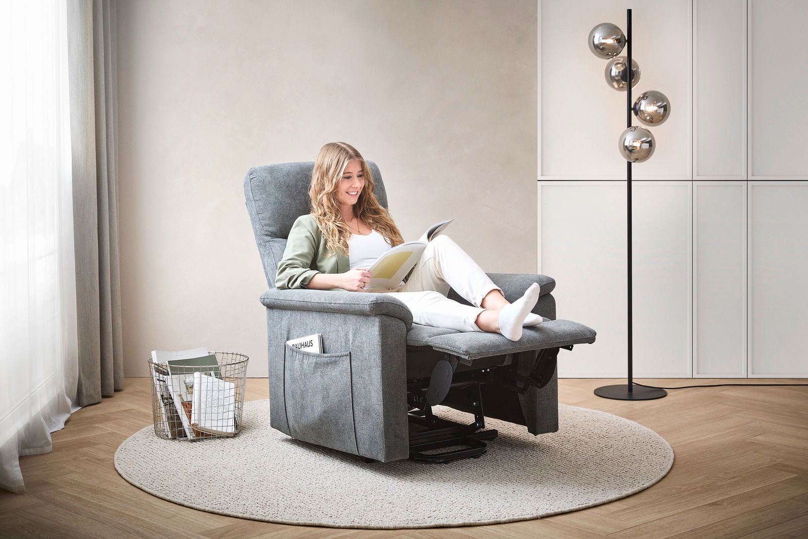 Frau entspannt in einem grauen Relaxsessel, seitliche Perspektive, mit einem Buch in der Hand, umgeben von einem modernen Wohnzimmerambiente.