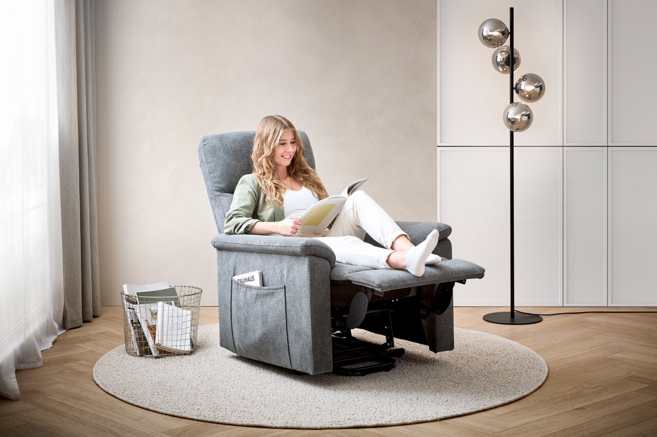 Frau entspannt in einem grauen Relaxsessel, seitliche Perspektive, mit einem Buch in der Hand, umgeben von einem modernen Wohnzimmerambiente.