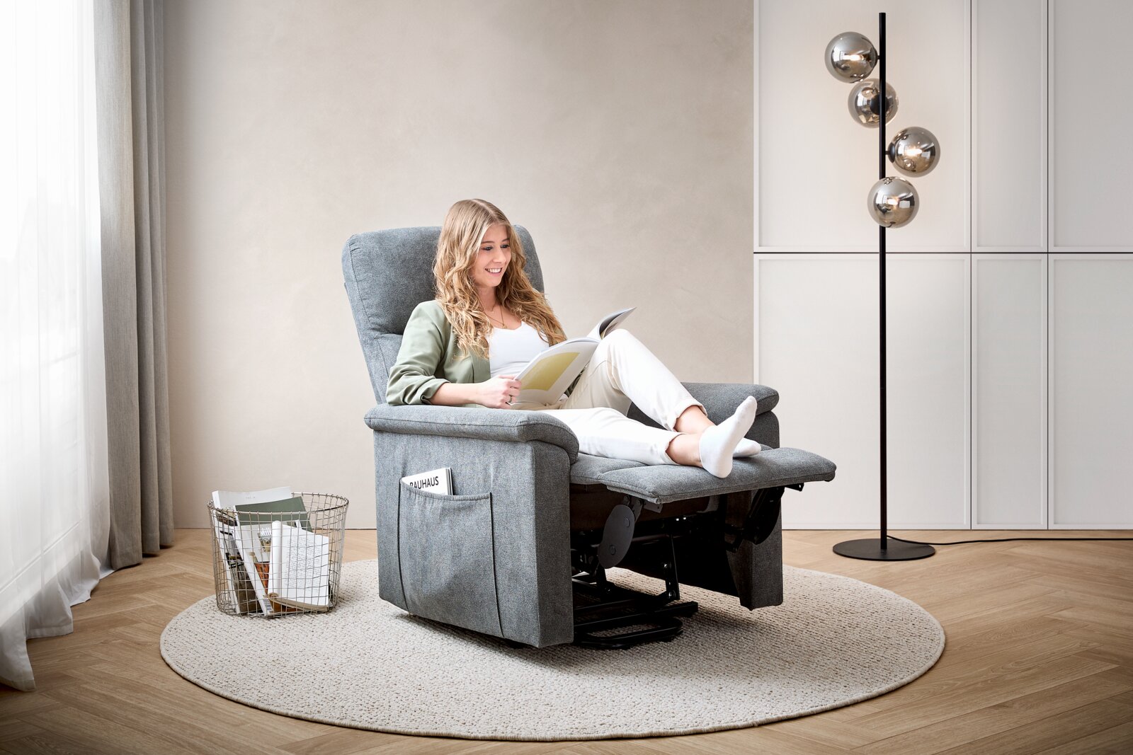 Frau entspannt in einem grauen Relaxsessel, seitliche Perspektive, mit einem Buch in der Hand, umgeben von einem modernen Wohnzimmerambiente.