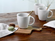 Weiße Keramiktasse mit Henkel und Löffel auf Holzbrett neben zwei Macarons auf rustikalem Holztisch, weitere Tassen unscharf im Hintergrund; Perspektive: leicht seitlich von vorn auf Tischhöhe.