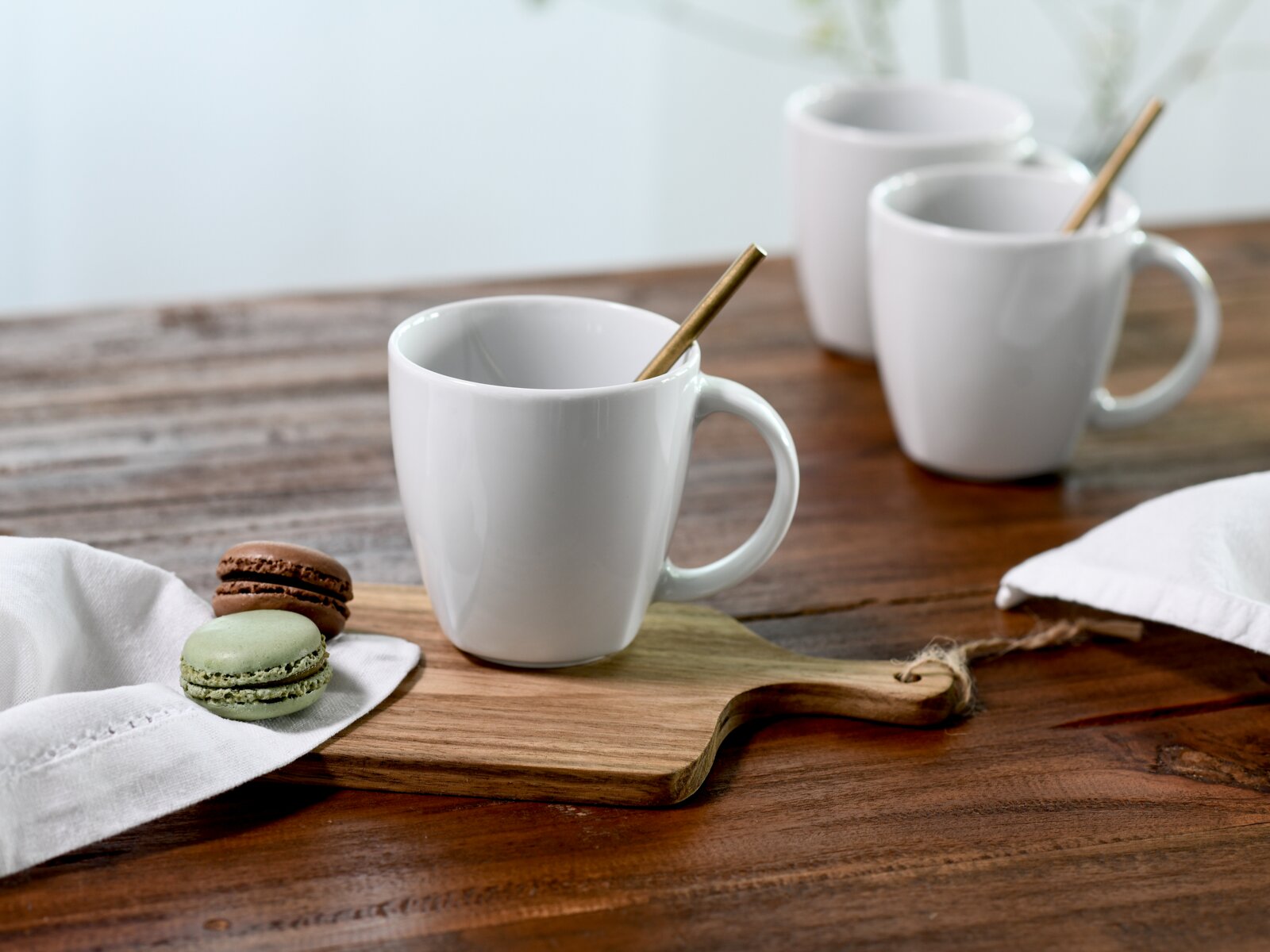 Weiße Keramiktasse mit Henkel und Löffel auf Holzbrett neben zwei Macarons auf rustikalem Holztisch, weitere Tassen unscharf im Hintergrund; Perspektive: leicht seitlich von vorn auf Tischhöhe.