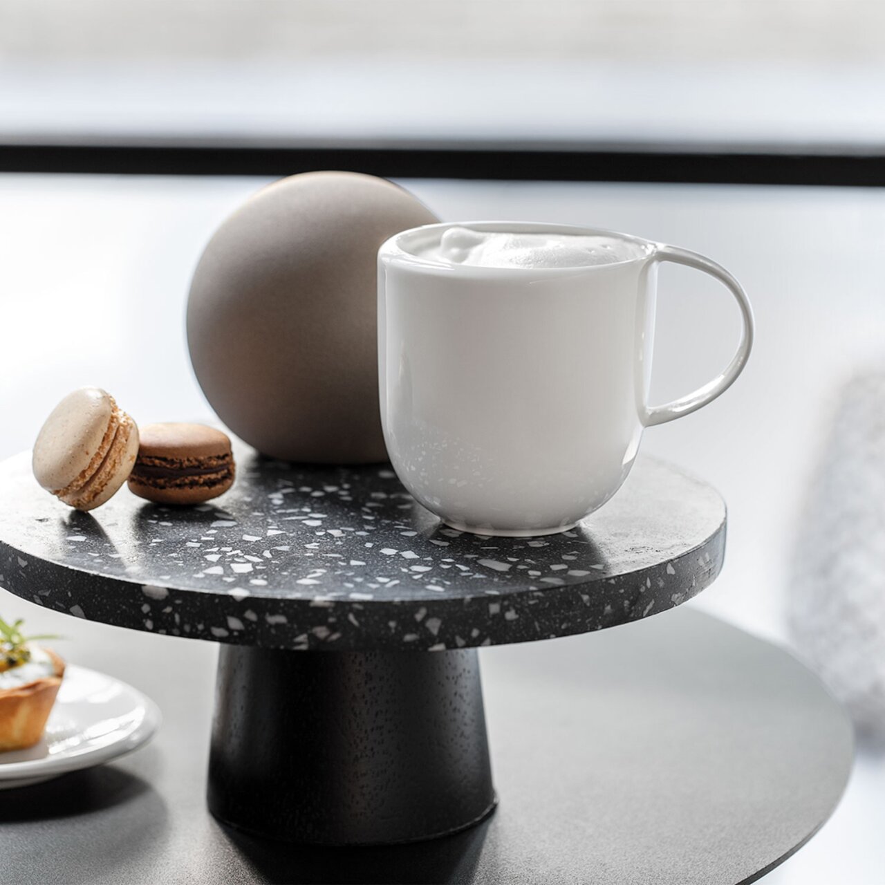 Villeroy & Boch Kaffeetasse NEW MOON Weißer Kaffeebecher NewMoon 310ml auf einem schwarzen Terrazzo-Tisch, seitliche Perspektive, mit Schaum gefüllt, neben Macarons und einer runden Dekoration.
