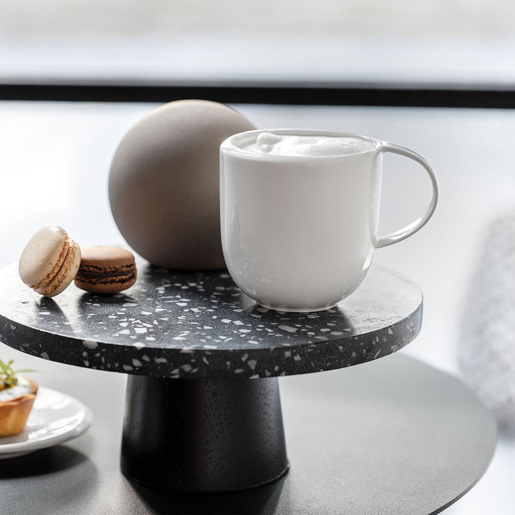 Weißer Kaffeebecher NewMoon 310ml auf einem schwarzen Terrazzo-Tisch, seitliche Perspektive, mit Schaum gefüllt, neben Macarons und einer runden Dekoration.
