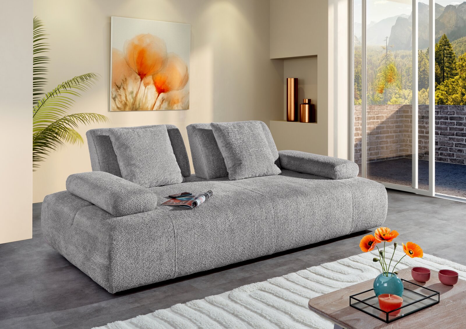 Graues 2-Sitzer Sofa in modernem Wohnzimmer, seitliche Perspektive, mit Kissen und Zeitschriften auf der Sitzfläche, umgeben von stilvoller Dekoration und großen Fenstern mit Blick auf die Natur.