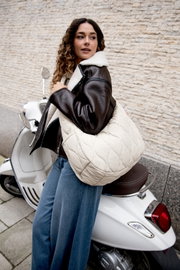 Een jonge vrouw draagt een grote crèmekleurige, gewatteerde totebag over haar schouder en zit op een witte scooter voor een stenen muur; zijaanzicht vanuit een licht onderhoek.