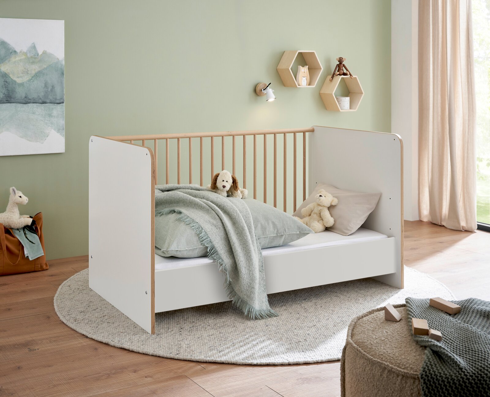 MyBaby Umbauseiten 2er Set ASTEA Modernes Babybett/Kinderbett in Weiß mit Holzrahmen und Gitterseiten, mit Bettwäsche und Kuscheltieren, im Kinderzimmer – Ansicht schräg von vorn.