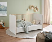 Modernes Babybett/Kinderbett in Weiß mit Holzrahmen und Gitterseiten, mit Bettwäsche und Kuscheltieren, im Kinderzimmer – Ansicht schräg von vorn.