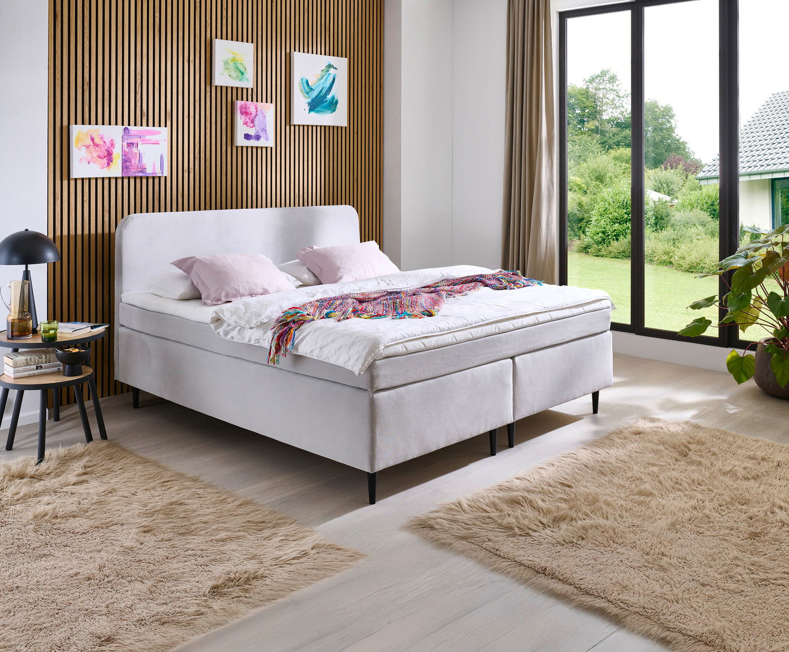 Modernes Boxspringbett in einem stilvollen Schlafzimmer mit Holzwandverkleidung und großen Fenstern, seitliche Perspektive.