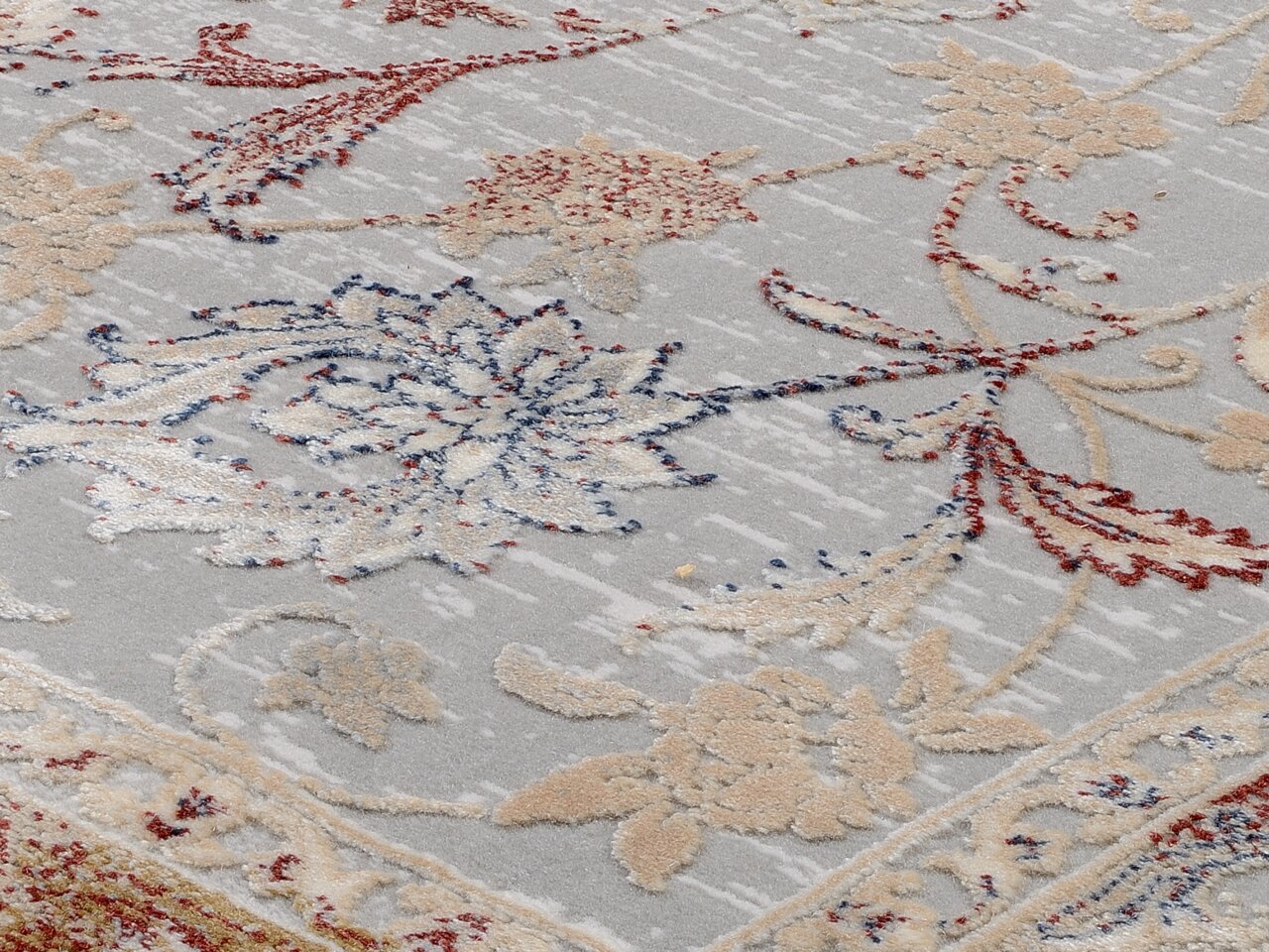Nahaufnahme eines Orientteppichs mit floralen Mustern in Rot, Blau und Beige auf grauem Hintergrund, perspektivisch von schräg oben aufgenommen.