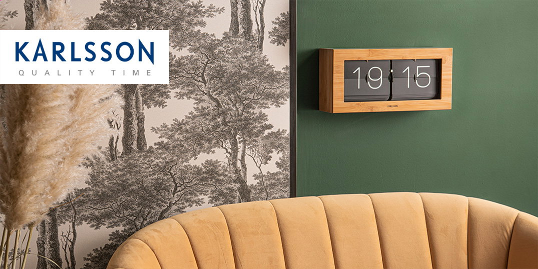 Een moderne wandklok met houten lijst hangt boven een gele fauteuil tegen een groene muur.