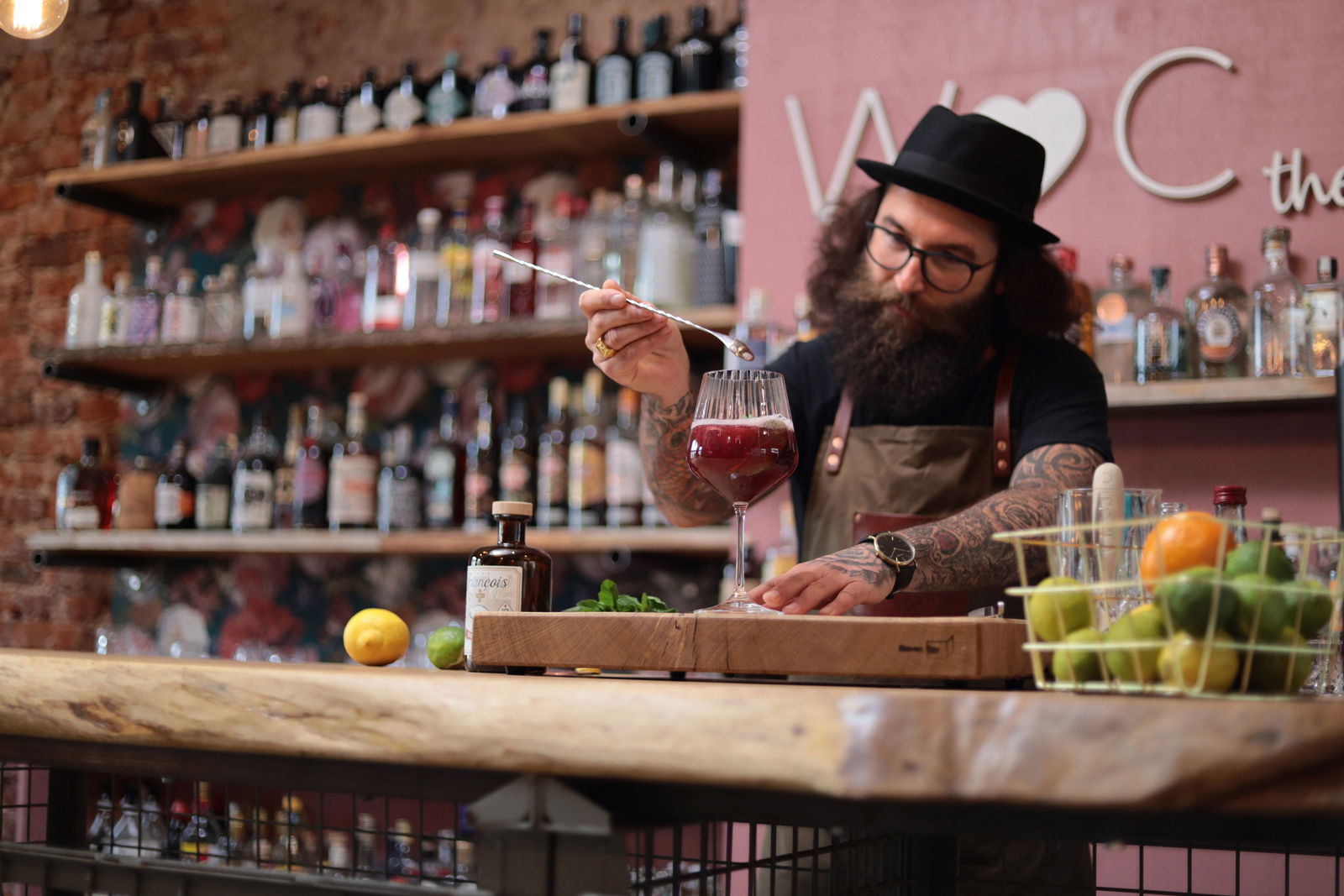 Ein Bartender mit Hut und Bart bereitet einen Drink in einem großen Weinglas zu. Im Hintergrund sind Regale mit verschiedenen Spirituosenflaschen zu sehen. Die Perspektive ist frontal auf den Bartender gerichtet.
