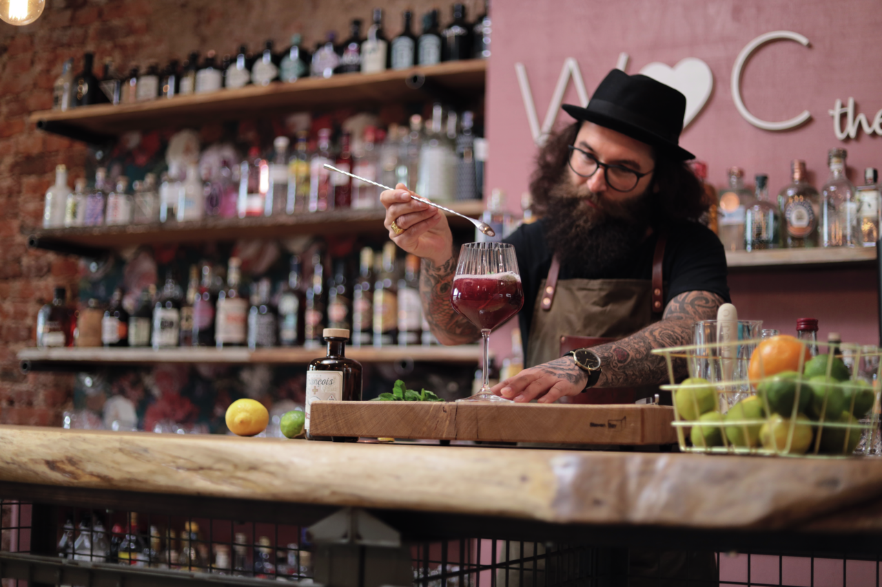 Nachtmann 4er-Set Gin Tonic-Gläser + Glas-Trinkhalme TASTES GOOD Ein Bartender mit Hut und Bart bereitet einen Drink in einem großen Weinglas zu. Im Hintergrund sind Regale mit verschiedenen Spirituosenflaschen zu sehen. Die Perspektive ist frontal auf den Bartender gerichtet.